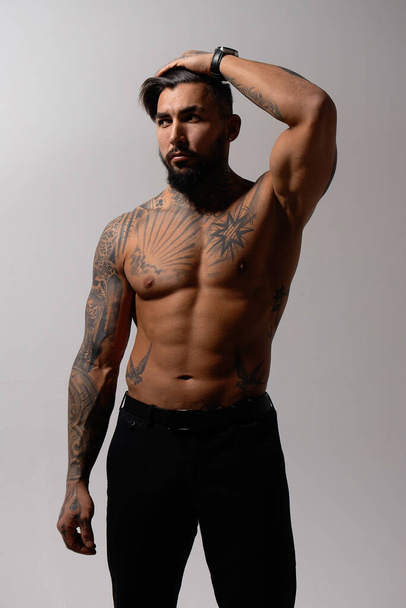 Ισπανικό ανδρικό μοντέλο χωρίς πουκάμισο με μυϊκό τατουάζ κορμό που στέκεται με τα χέρια στις τσέπες και κοιτάζει μακριά σε γκρι φόντο - Φωτογραφία, εικόνα