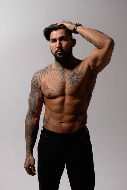 Hispanic shirtless male model with muscular tattooed torso standing with hands in pockets and looking away on gray backdrop - Foto, imagen