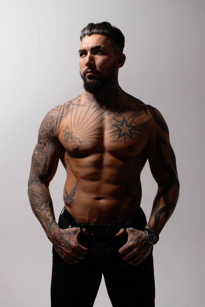 Modelo masculino hispano sin camisa con torso musculoso tatuado de pie con las manos en los bolsillos y mirando hacia otro lado sobre fondo gris - Foto, Imagen