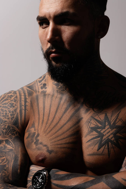 Modèle masculin sans chemise hispanique avec torse tatoué musclé debout avec les mains dans les poches et regardant loin sur fond gris - Photo, image