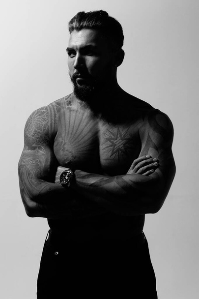 Іспаномовна модель чоловіка з м'язистим татуюванням тулуба стоїть з руками в кишенях і дивиться на сірий фон. - Фото, зображення