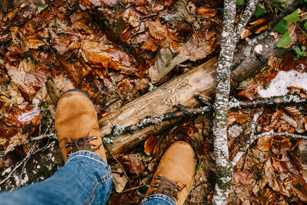 Patas masculinas en botas marrones sobre hojas caídas junto a un enganche de madera. Primer plano - Foto, imagen