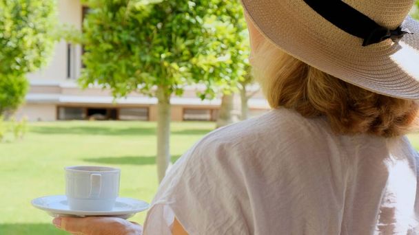 Senora femmina, 50-55 anni in un cappello di paglia tiene una tazza di bevanda calda e beve caffè, seduta al mattino su una terrazza estiva in una giornata di sole. Il concetto di buongiorno, viaggio e aspettativa di - Foto, immagini