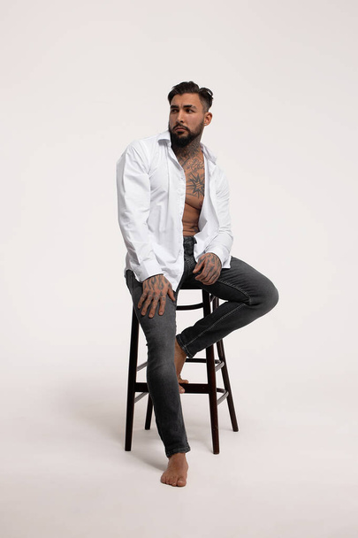 Hispanic shirtless male model with muscular tattooed torso standing with hands in pockets and looking away on gray backdrop - Foto, Imagem