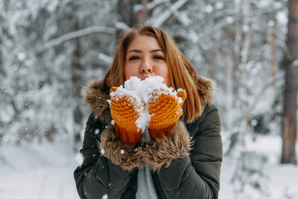 A happy young girl blows on the white snow, which lies on cozy knitted woolen mustard-colored mittens. Blurred snowflakes in the foreground. Christmas holidays, Winter time. without a face. - Foto, Imagen