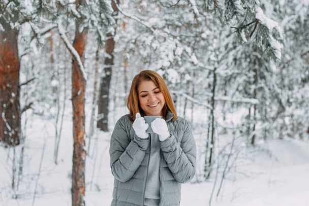 A cute girl with short hair in a gray jacket and knitted white mittens is drinking hot tea in a snowy forest. Cozy and warm in the winter season. - Foto, imagen
