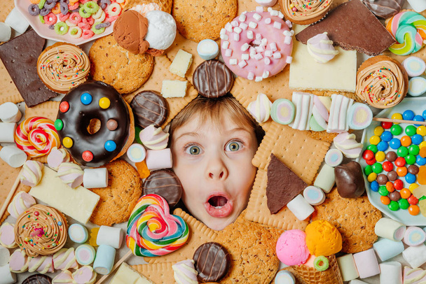 Funny 5 years old girl lying under plenty of sweet foods. Kids face surrounded by unhealthy food. Top view, flat lay. - Фото, изображение