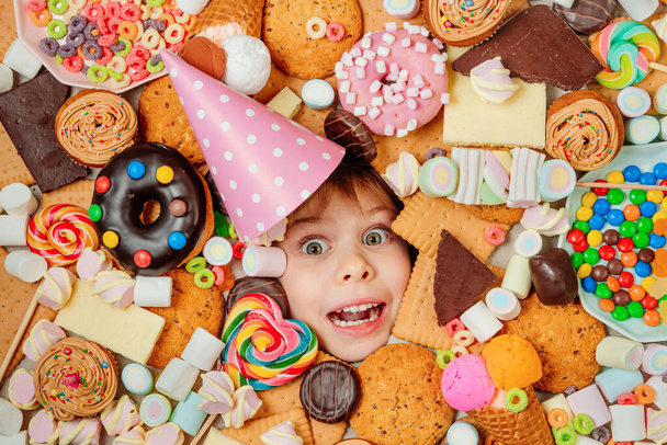 Joyeux 5 ans fille portant chapeau d'anniversaire couché sous beaucoup d'aliments sucrés. Les enfants sont entourés d'aliments malsains. Concept de fête pour enfants. Vue de dessus, plan plat. - Photo, image