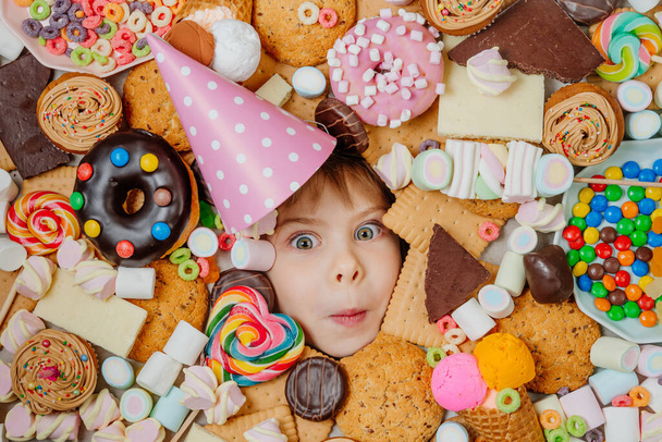 Feliz niña de 5 años con sombrero de cumpleaños acostado debajo de un montón de alimentos dulces. Los niños se enfrentan rodeados de comida poco saludable. Concepto de fiesta infantil. Vista superior, plano. - Foto, imagen