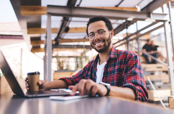 Portrait of cheerful man in optical glasses for provide eyes correction smiling at camera while touching smartphone screen during coffee break in coworking space for freelancers, technology concept - Photo, image