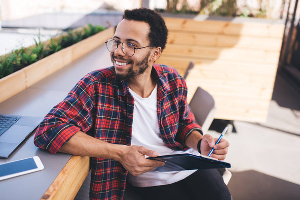 Happy male designer in classic spectacles for vision protection enjoying creative paperwork, cheerful Hispanic student in eyewear looking away and smiling while learning and studying indoors - Photo, image