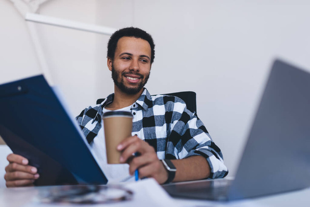 Cheerful employer watching informative webinar while planning business organization in office interior, happy male professional with paperwork and coffee to go browsing website on laptop technology - Photo, Image