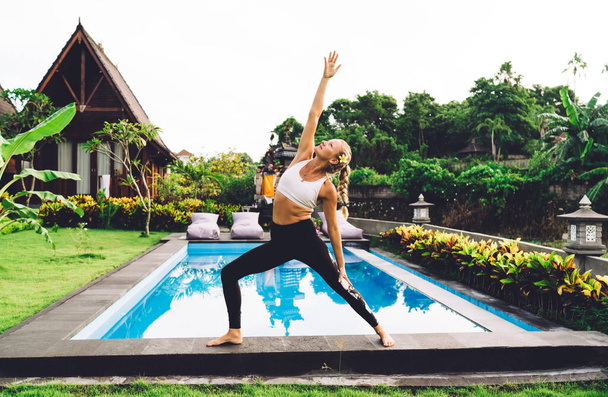 Side view of energetic fit female in activewear practicing yoga and standing in warrior pose with hand raised up near swimming pool - Foto, afbeelding