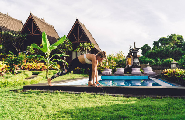 Side view of concentrated female practicing yoga and standing in Warrior pose while spending time near swimming pool in tropical resort on sunny day - Zdjęcie, obraz