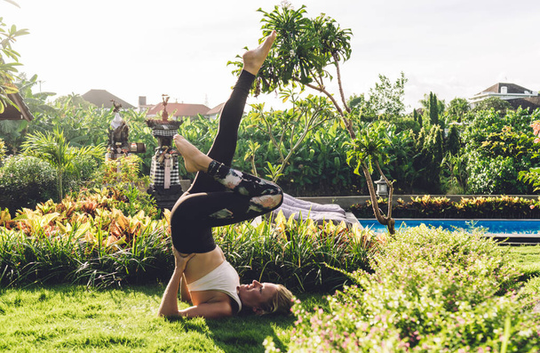 Side view of female in white top and black leggings doing yoga in Supported Shoulder Stand pose on sunny summer day on tropical resort - Foto, Bild