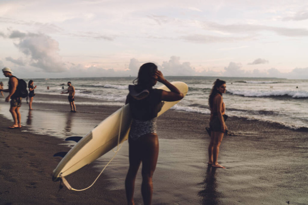 Back view of anonymous female in swimwear holding surfboard while standing on sandy coast crowded with people near waving sea - Foto, Imagen