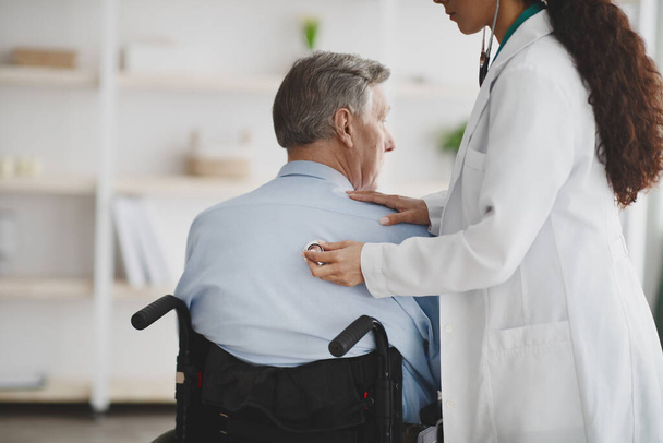 Female doctor using stethoscope on older man in wheelchair at home - Foto, imagen