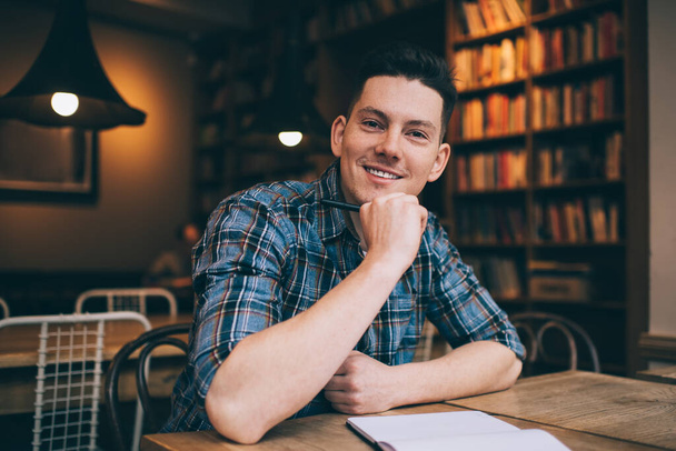 Cheerful young man with pen in hand touching chin while sitting in library and smiling at camera in soft lamp light - Photo, Image