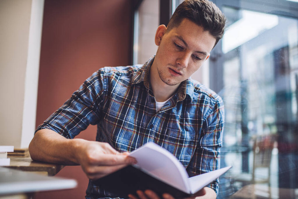 Intelligent male student reading informative notes during time for knowledge studying and exam preparation, serious man 20 years old analyzing textbook info while doing university homework - Photo, image