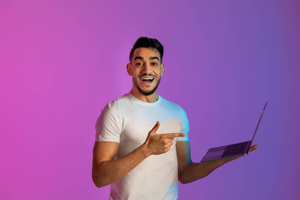 Iloinen nuori arabimies osoittaa kannettavaan tietokoneeseen, toimii verkossa, kommunikoi verkossa neonvalolla - Valokuva, kuva