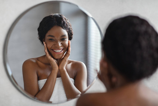 Αντανάκλαση καθρέφτη της νεαρής μαύρης γυναίκας που εφαρμόζει προϊόν προσώπου - Φωτογραφία, εικόνα