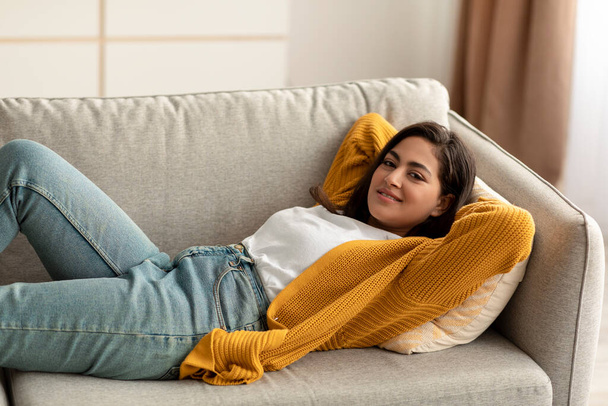 Du calme. Joyeux arabe femme couché avec les mains derrière la tête sur un canapé confortable et souriant, avoir une grande journée de congé - Photo, image