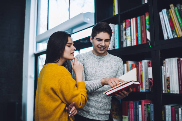 Young positive man in casual wear pointing at article in opened book and showing concentrated girlfriend while standing together against bookshelf - Фото, изображение