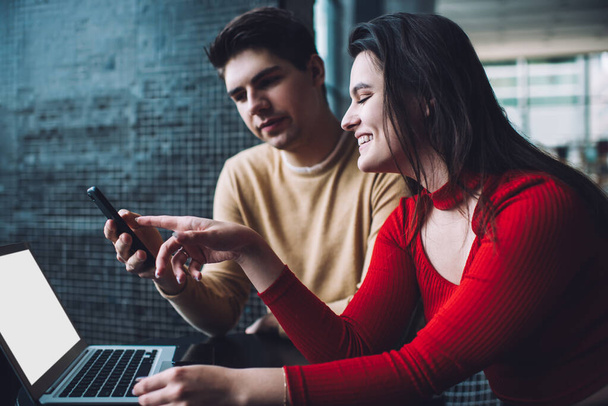 Content smiling young woman in red casual clothes using smartphone with male colleague and pointing at screen while working together on netbook with blank screen in creative workplace - Фото, изображение