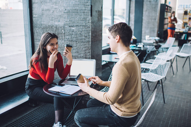 Side view of young man and woman in casual clothes sitting at table with laptop and documents while discussing details of project in contemporary cafe - Foto, afbeelding