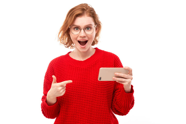 Shocked joyful redhead girl points finger at smartphone blank screen in spectacles and red clothes isolated on white studio background - Zdjęcie, obraz