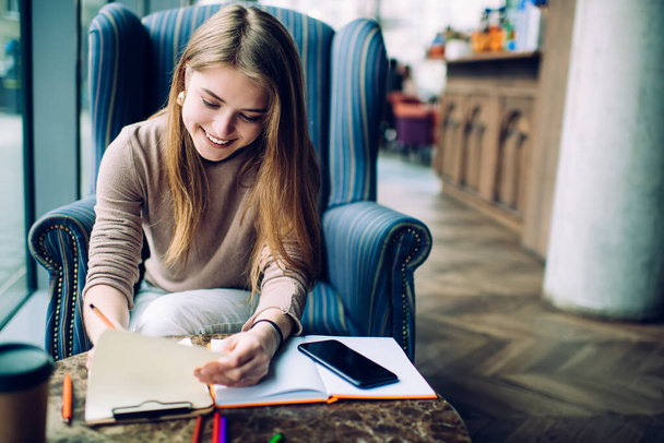 Cheerful female freelancer in casual outfit sitting in comfortable armchair and taking notes on clipboard while smiling in cozy cafe - Foto, Imagen