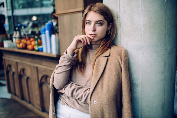 Charming female in beige sweater with hand on chin standing near counter and looking wistfully away during break from work process - Photo, Image
