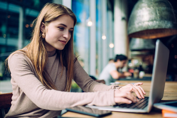 Positive young long haired woman in casual wear typing on keyboard of modern laptop while working alone in cozy cafe - Fotoğraf, Görsel