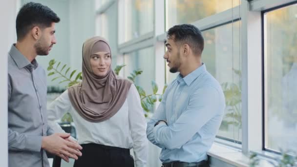 Tres colegas profesionales diversos que hablan en la oficina moderna, mujer musulmana árabe amistosa feliz en hijab y dos compañeros de trabajo indios de los hombres que tienen conversación discuten proyecto de negocio en el espacio de trabajo - Imágenes, Vídeo