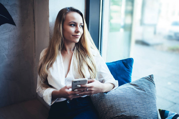 Happy young long haired female in trendy clothes sitting on windowsill with cushions and messaging using mobile phone in apartment - Foto, afbeelding