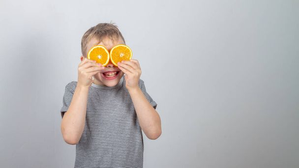 A Caucasian boy in a gray T-shirt on a gray background in the studio closed his eyes with orange orange slices and laughs happily. - Фото, изображение