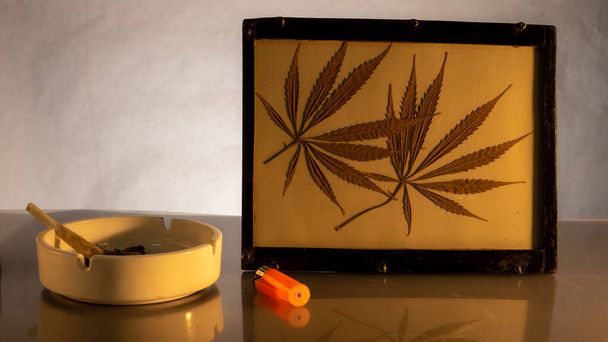 Kurutulmuş marihuana yapraklı bir resim, kül tablasının yanında elle sarılmış bir puro. - Fotoğraf, Görsel