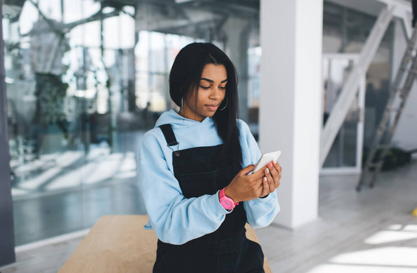 Black girl watching something on mobile phone while standing by wooden table. Concept of modern woman lifestyle. Young female person wearing casual clothes and wristwatch. Blurred interior of office - Fotó, kép