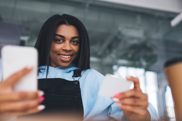 Smiling african girl holding business card and mobile phone. Concept of modern woman lifestyle. Selective focus of young female person wearing casual clothes. Lady looking at camera - Photo, image