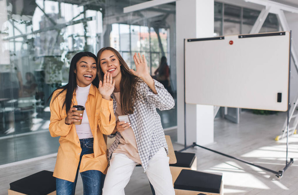 Smiling businesswomen waving hands at somewhere in office. Concept of modern successful women. Idea of friendship. Black girl with coffee cup. European lady with mobile phone. Girls sitting on desk - Photo, Image