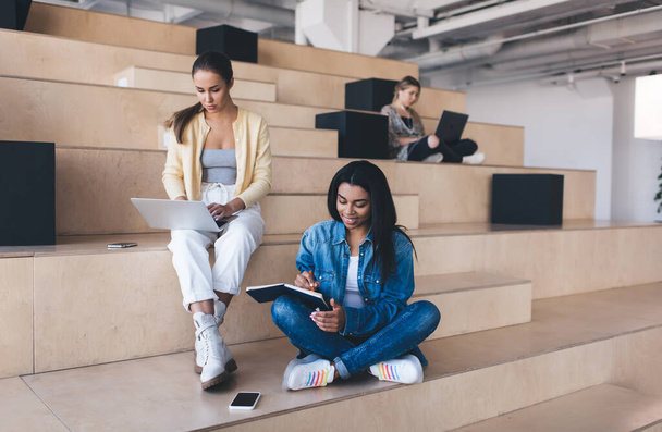Businesswomen working on wooden benches in coworking office space. European woman using laptop computer. Smiling black girl writing something in notebook. Concept of modern successful women - Photo, Image