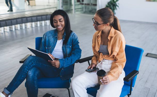 Black businesswoman showing something in notebook to european female colleague with coffee and mobile phone. Concept of modern successful women. Idea of teamwork. Young women sit in chairs in office - Foto, imagen