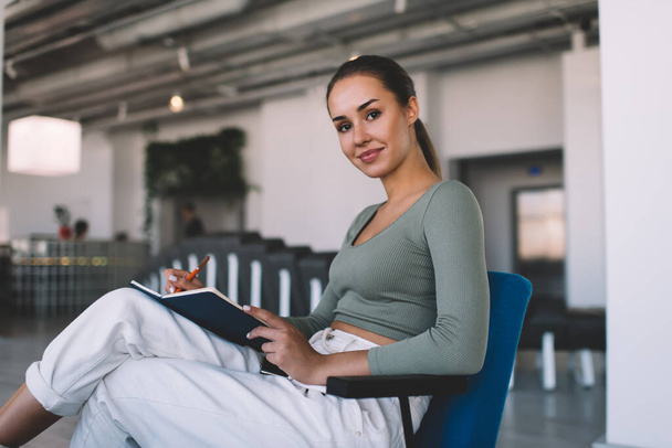 European businesswoman writing something in notebook with pen during work. Concept of modern successful woman. Young smiling female person sitting in chair and looking at camera. Interior of office - Photo, image
