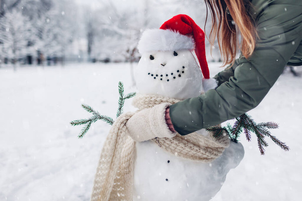 Девушка делает снеговика на свежем воздухе в снежном зимнем парке, одевая его в шляпу Санты, шарф и прикрепляя елки в руки. Рождественские развлечения - Фото, изображение