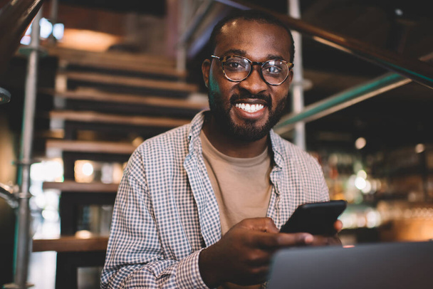 Portrait of cheerful male blogger with cellphone gadget and cropped laptop smiling at camera during free time, happy African American man in classic glasses using smartphone and netbook indoors - Foto, Bild