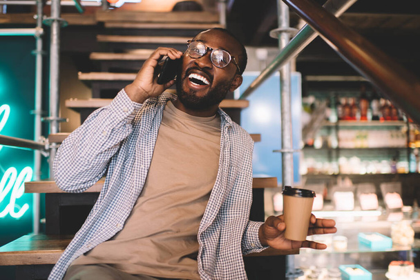 Excited male in classic spectacles for provide vision correction laughing during friendly cellular conversation, funny dark skinned man with takeaway coffee to go rejoicing during mobile talk - Foto, Imagen