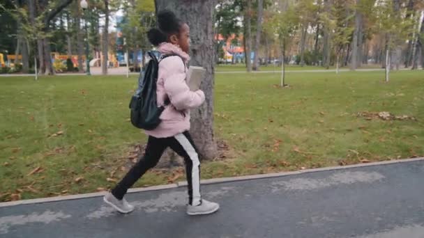 Side view small schoolgirl little afro american girl child african baby pupil with backpack and book walking street in park back to school. Alone cheerful kid walks going outdoors in autumn background - Footage, Video