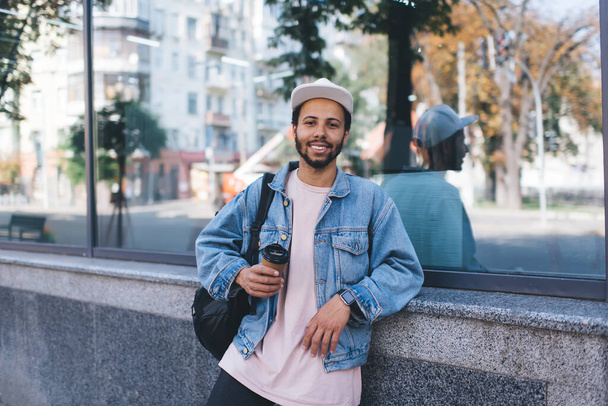 Half length portrait of carefree hipster guy in trendy denim clothes smiling at camera during coffee break in touristic city, happy traveller in cap holding takeaway caffeine beverage and posing - Фото, изображение