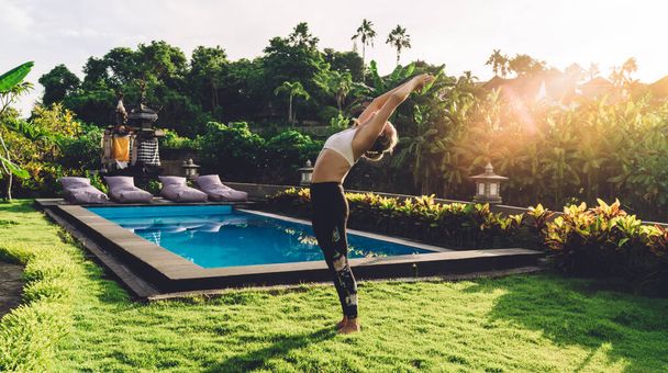 Unrecognizable female stretching body in green park and doing breathing exercises while enjoying sunny day against swimming pool during countryside holidays - Photo, image