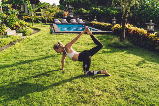 From above of slim female athlete doing yoga asana on grass near blooming bushes and swimming pool during summer holidays - Photo, Image